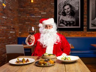 A Person In A Santa Suit Sitting At A Table With Food And A Glass Of Wine